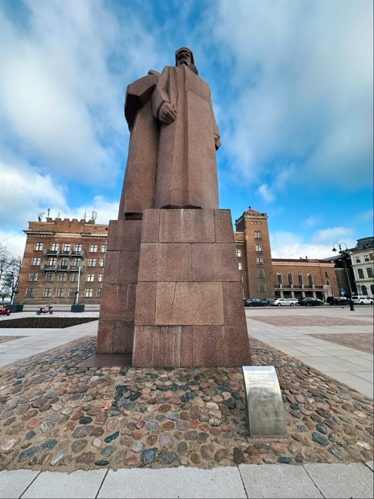 A towering reddish-brown stone statue of a man on a high pedestal in Riga surrounded by cobblestones and brick buildings