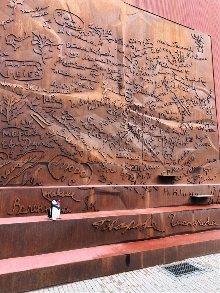 A large rusted metal wall covered in engraved signatures and handwriting with Miško the knitted penguin sitting on a ledge in Riga, Latvia