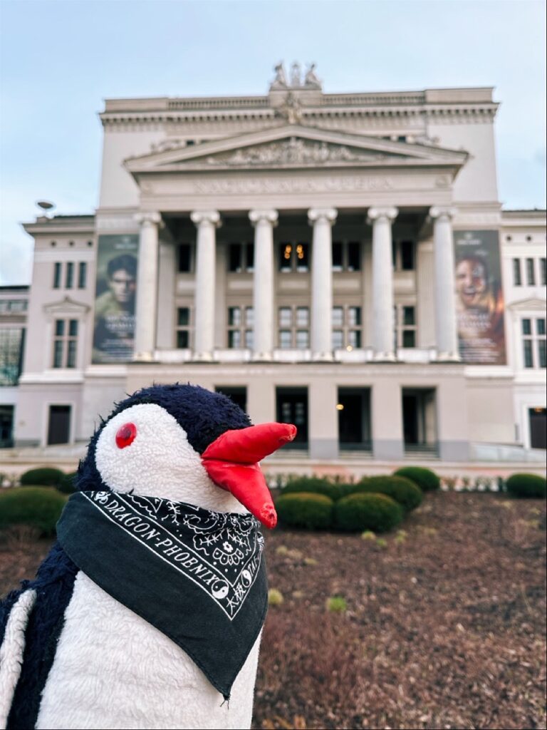 Miško the knitted penguin wearing a black bandana with Dragon Phoenix text stands before a large white building with columns in Riga