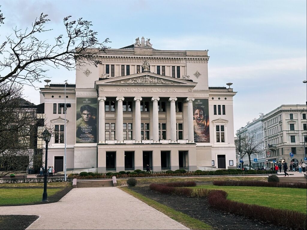 The white neoclassical facade of the building labeled NACIONĀLĀ OPERA in Riga with banners and people walking in the park