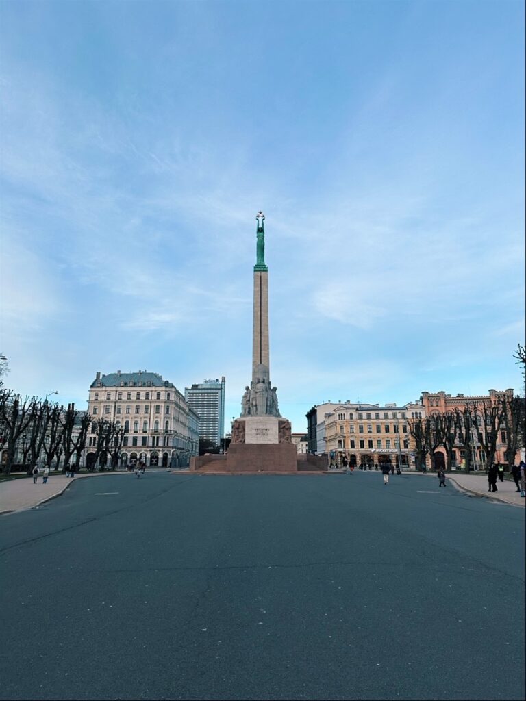 A tall monument topped with a green statue holding stars stands in a wide square in Riga surrounded by buildings and people walking