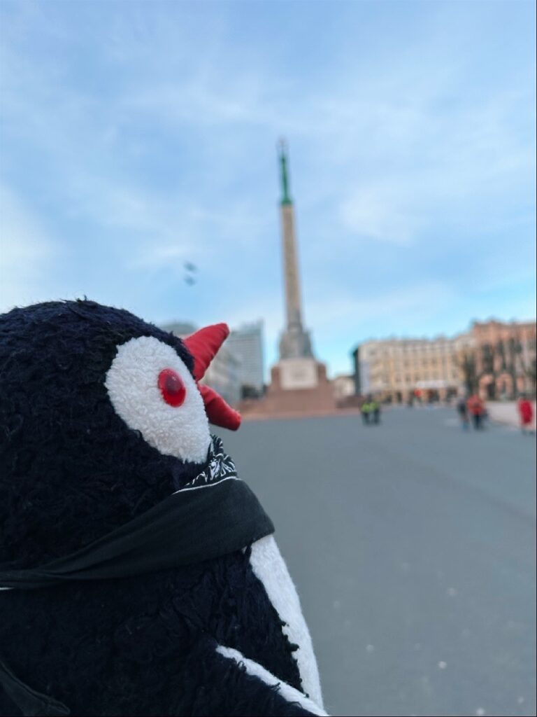 Miško the knitted penguin with a red beak looks towards a tall monument in a square in Riga, Latvia