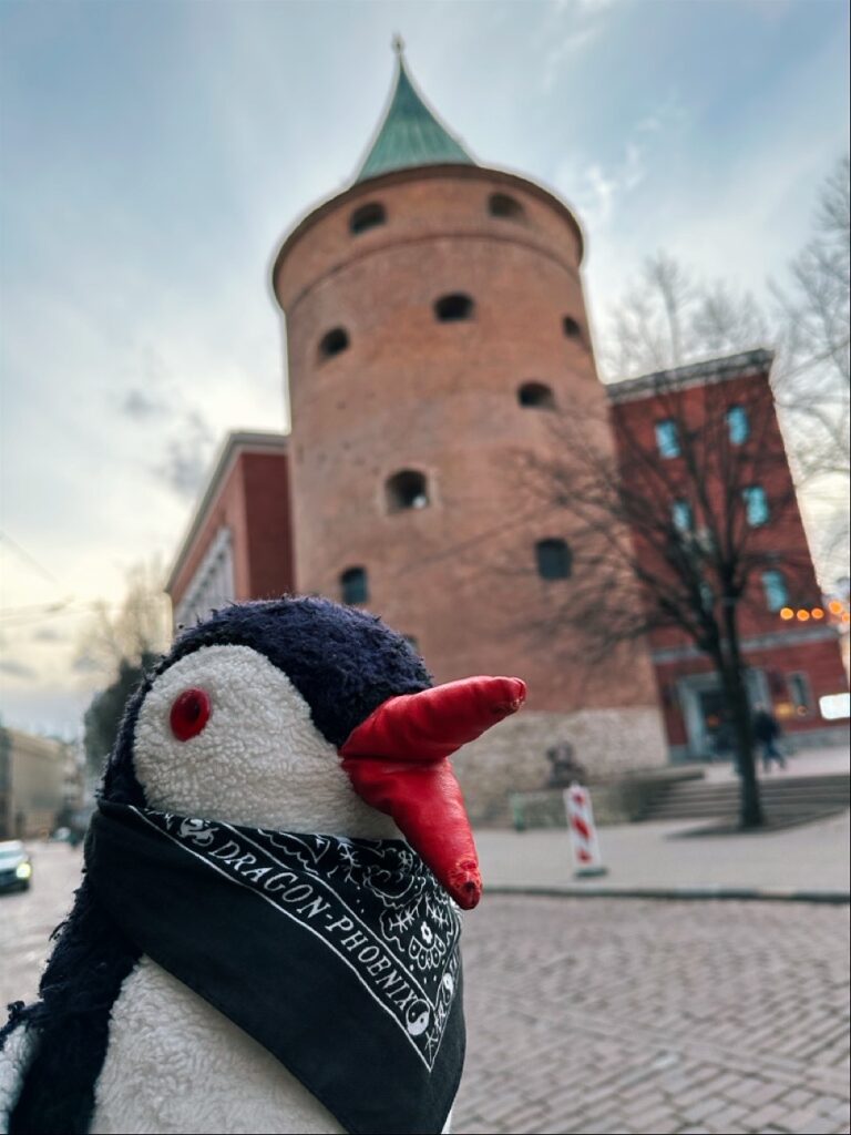 Miško the knitted penguin wearing a black Dragon-Phoenix bandana stands before a round brick tower with a green roof in Riga
