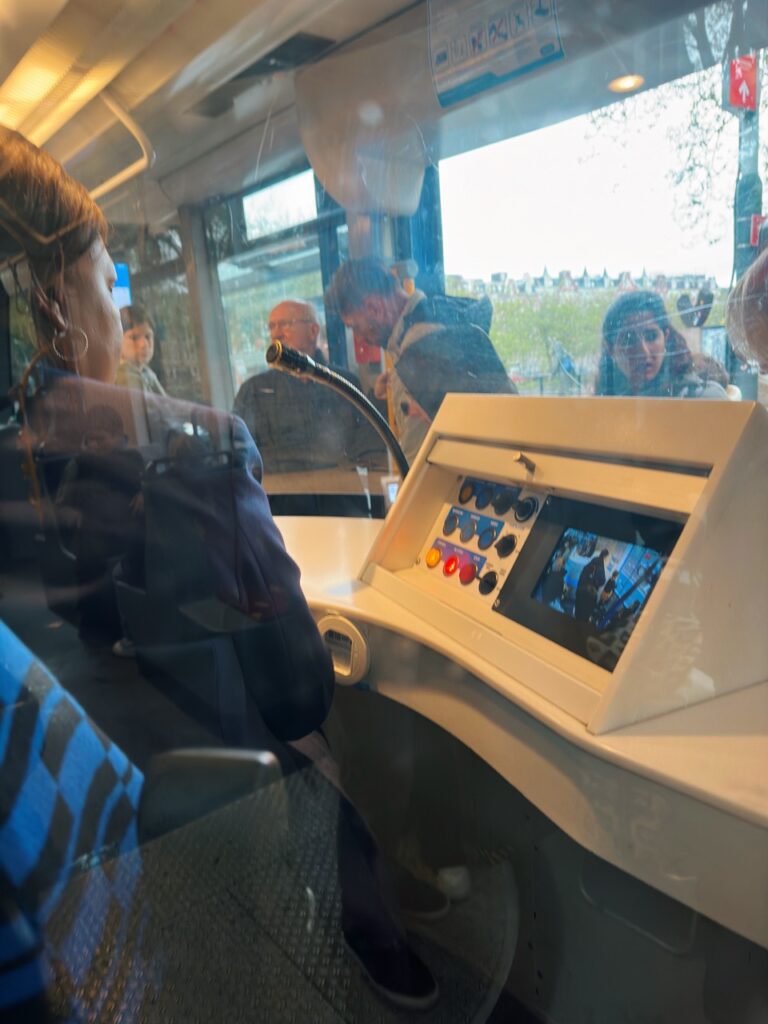 View inside an Amsterdam tram showing the driver's control console with buttons and a screen with passengers visible through the glass