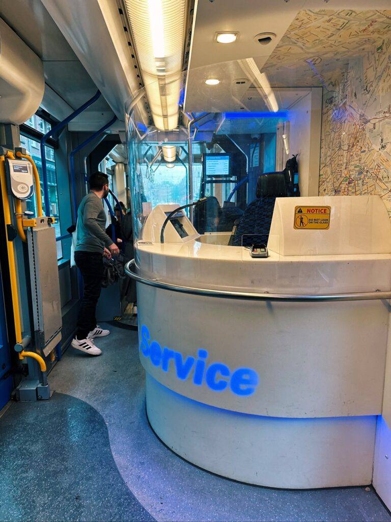 Interior of an Amsterdam tram showing a curved white service desk with blue Service text and a man standing near the yellow handrails