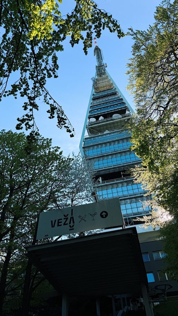 Low angle shot of the glass VEŽA tower framed by trees with a sign below reading VEŽA and symbols for a restaurant and bar
