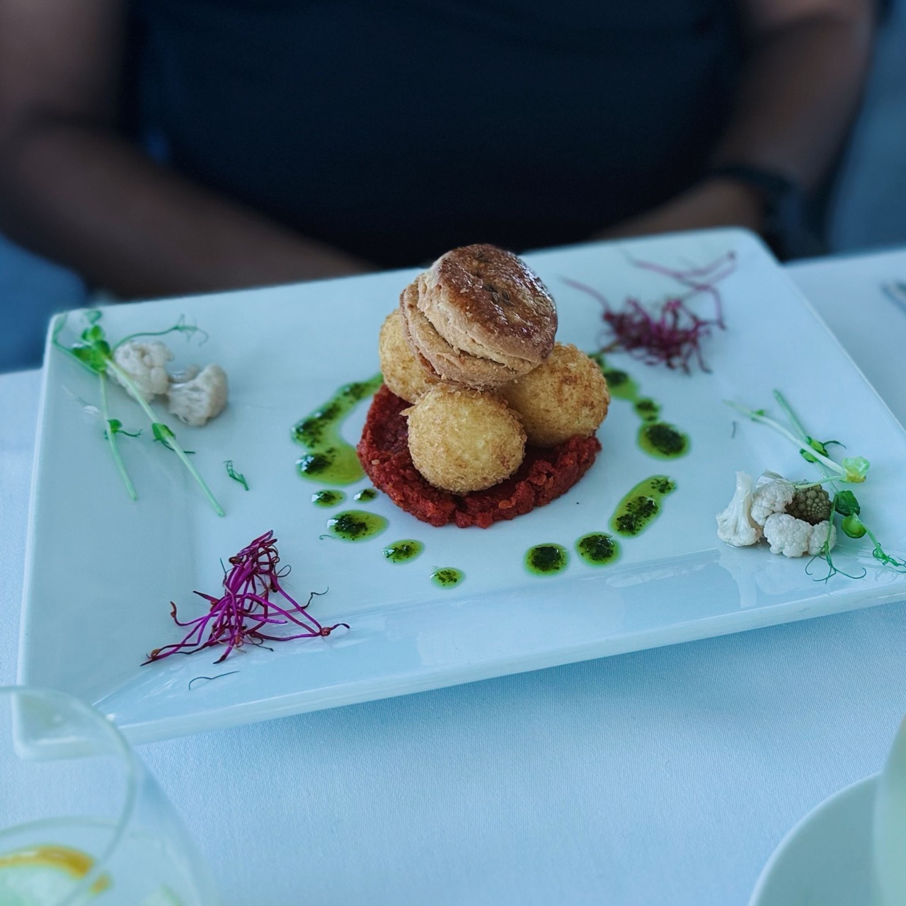 Stacked fried balls on a red base topped with a pastry, garnished with green oil and purple sprouts on a white rectangular plate