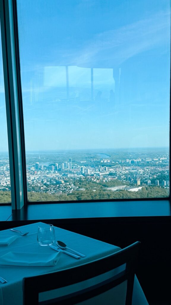 A dining table set with a white cloth and cutlery overlooks the city of Bratislava through a large window at VEŽA Bratislava