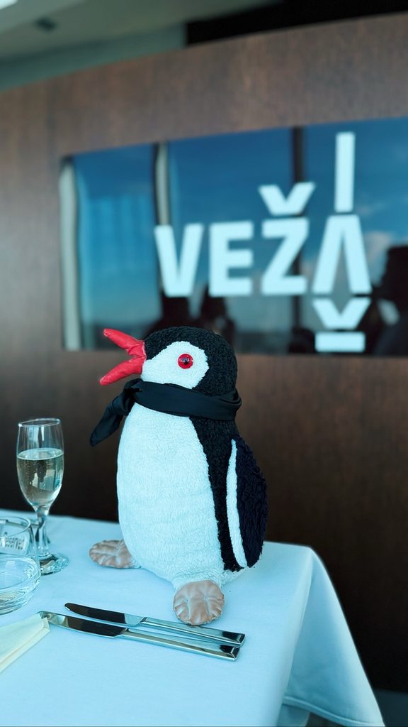Miško the knitted penguin sits on a white tablecloth with cutlery and a glass in front of a window sign reading VEŽA