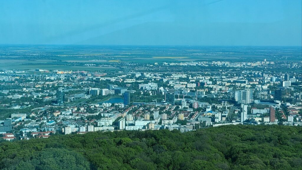 Panoramic view of Bratislava showing the city skyline and flat plains beyond a foreground of green trees