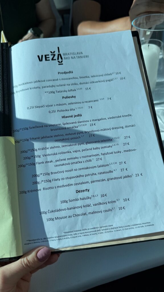 The menu for VEŽA Bratislava held by a hand listing sections for Predjedlá, Polievky, Hlavné jedlá and Dezerty with prices