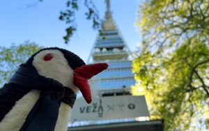 A knitted penguin with a red beak and black scarf looks up at the VEŽA tower sign against a blue sky and green trees