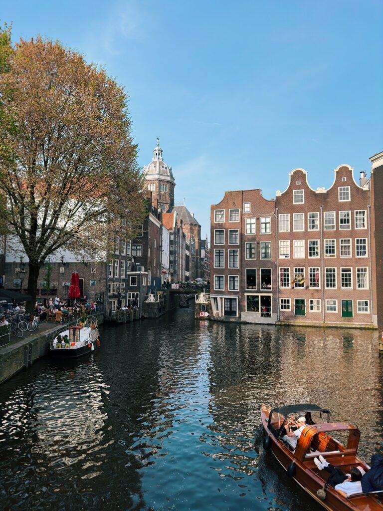 Wooden tour boat with passengers cruising an Amsterdam canal past traditional gabled brick buildings and a large tree