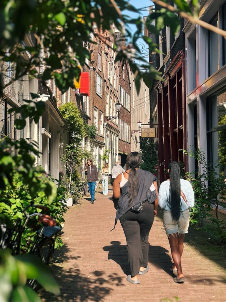 Two women walking away down a narrow brick street in Amsterdam lined with tall buildings and lush greenery