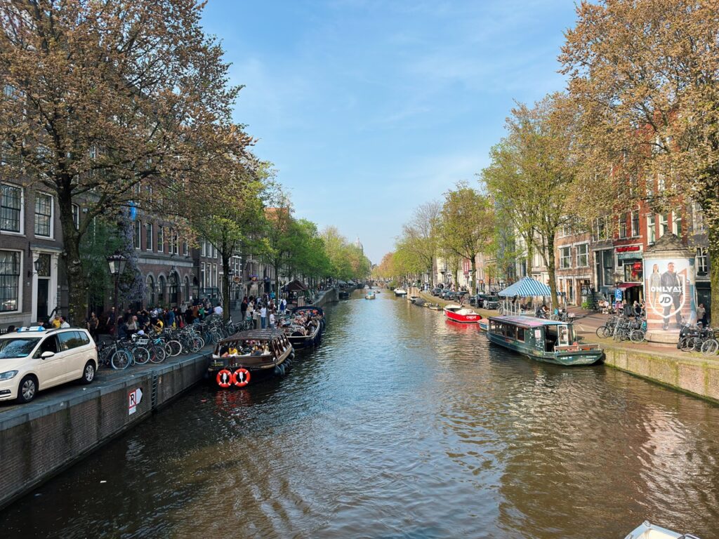 Amsterdam canal with brick buildings, trees, parked bicycles, and tour boats floating on the water under a blue sky