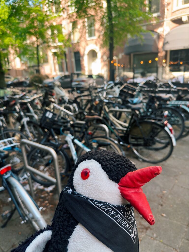Miško the knitted penguin wearing a black bandana stands in front of rows of parked bicycles in Amsterdam