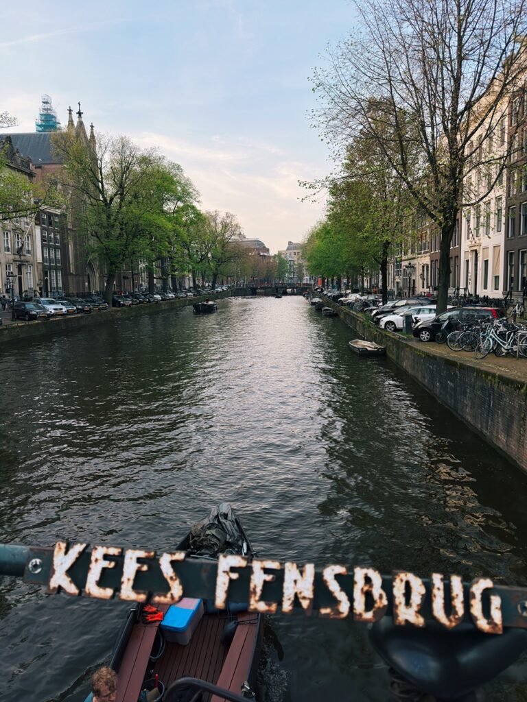 Rusty bridge sign reading KEES FENSBRUG overlooking an Amsterdam canal lined with green trees and parked cars on both sides