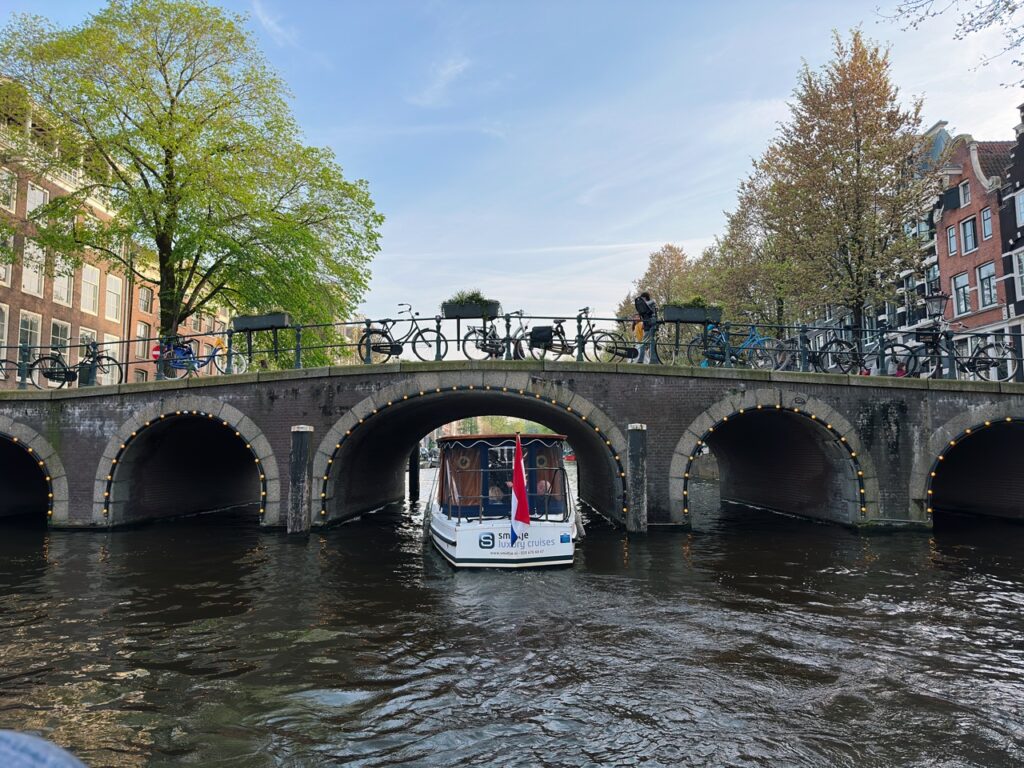 A white tour boat passes under a multi-arched brick bridge lined with bicycles in Amsterdam
