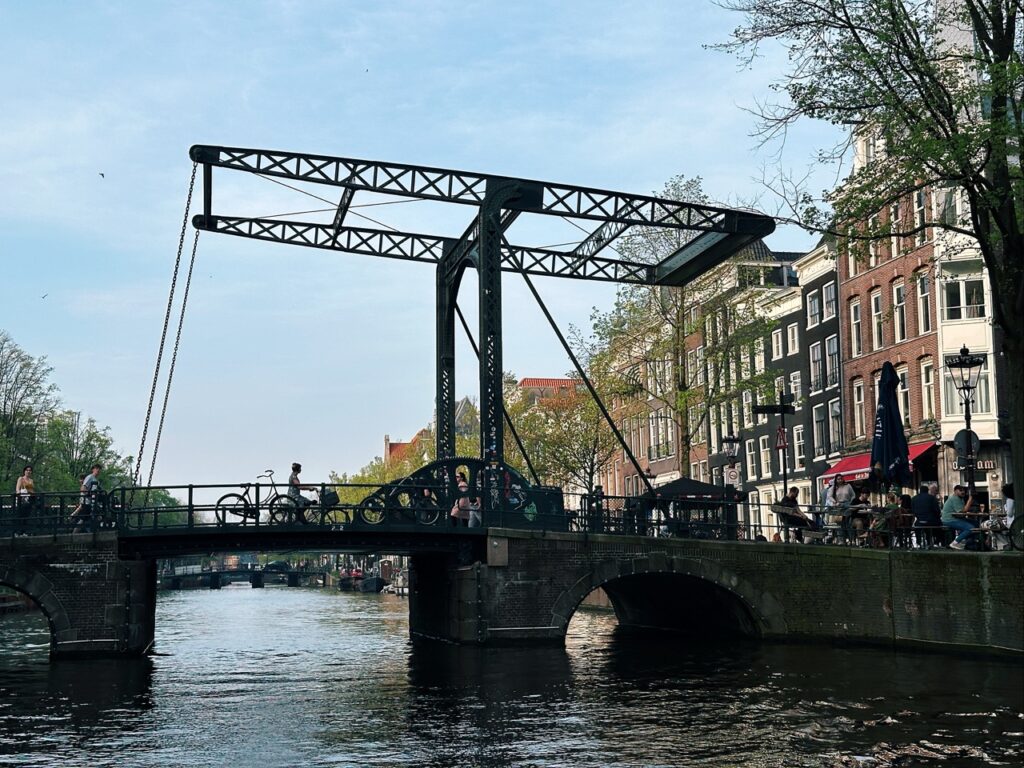 A black metal bridge over a canal in Amsterdam with cyclists crossing and people at outdoor tables near brick buildings