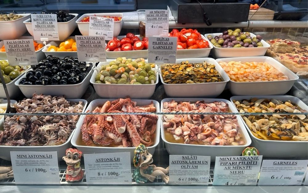 A market display of filled olives and mixed-cheese pastries, with Latvian menu text listing various fillings