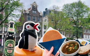 Miško the knitted penguin poses with a Heineken beer, a stroopwafel, and soup in front of Amsterdam canal houses
