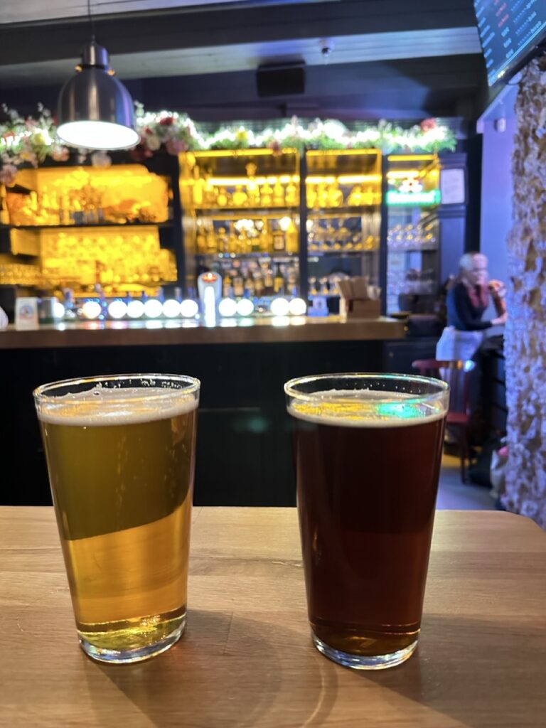 Two glasses of beer, one light and one dark, rest on a wooden table before a bar counter with beer taps and a woman seated