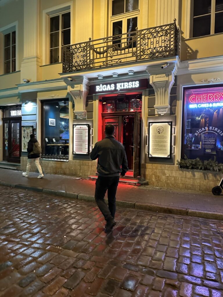 A man walks towards the entrance of a restaurant with a sign reading RĪGAS ĶIRSIS on a wet cobblestone street at night