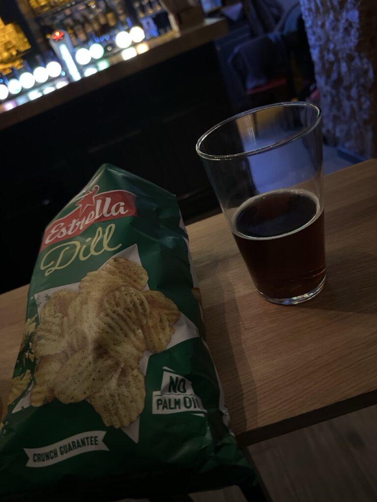 A green bag of Estrella Dill chips sits next to a glass of dark beer on a wooden table