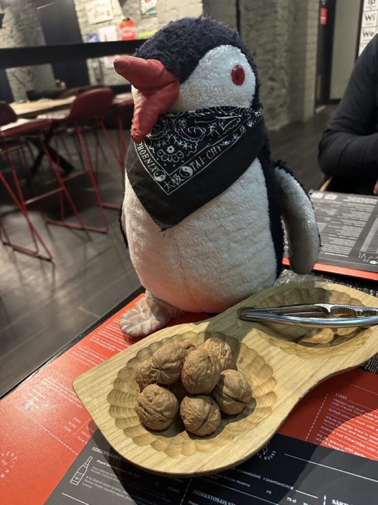 Miško the knitted penguin plush standing beside a restaurant sign