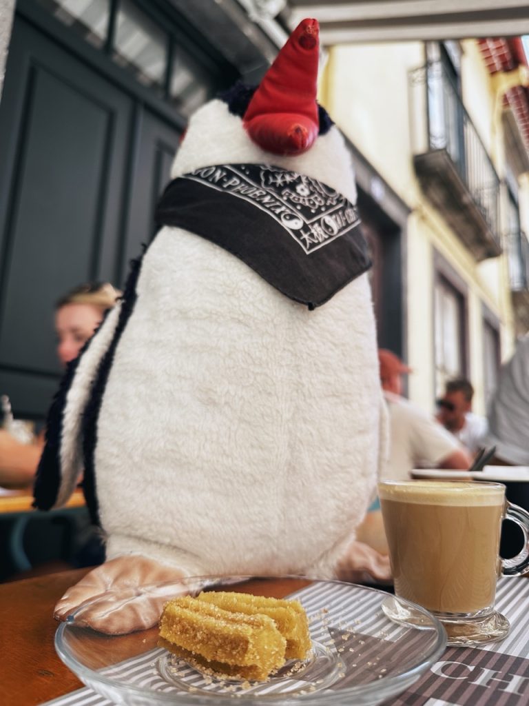 A plush penguin wearing a bandana sits beside coffee and churros in Madeira