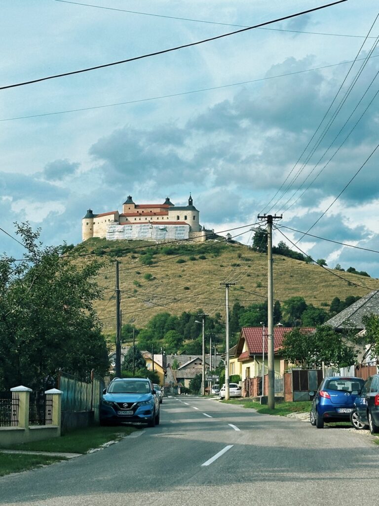 A large castle sits atop a grassy hill overlooking a quiet village street