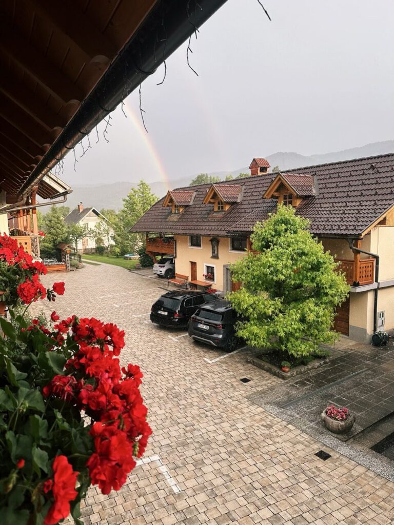 Rainbow arches over a courtyard with parked cars and red flowers in Slovenia