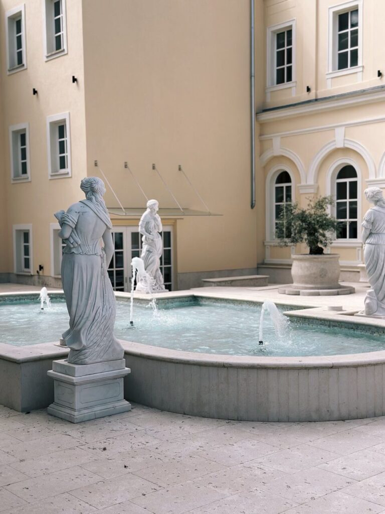 Classical statues stand around a stone fountain in a quiet courtyard in Piran