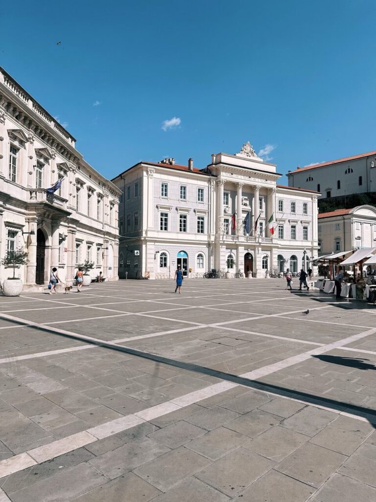People walk across a sunny stone square in Piran surrounded by historic buildings