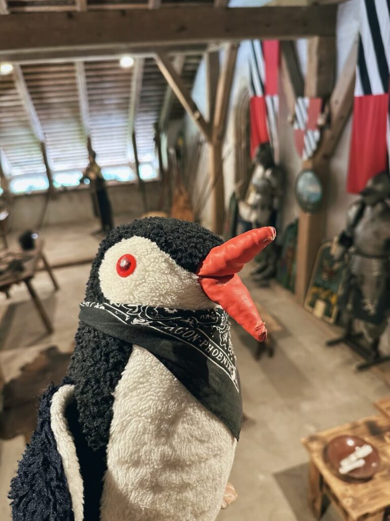 Plush penguin wearing a bandana inside a room with medieval armor displays