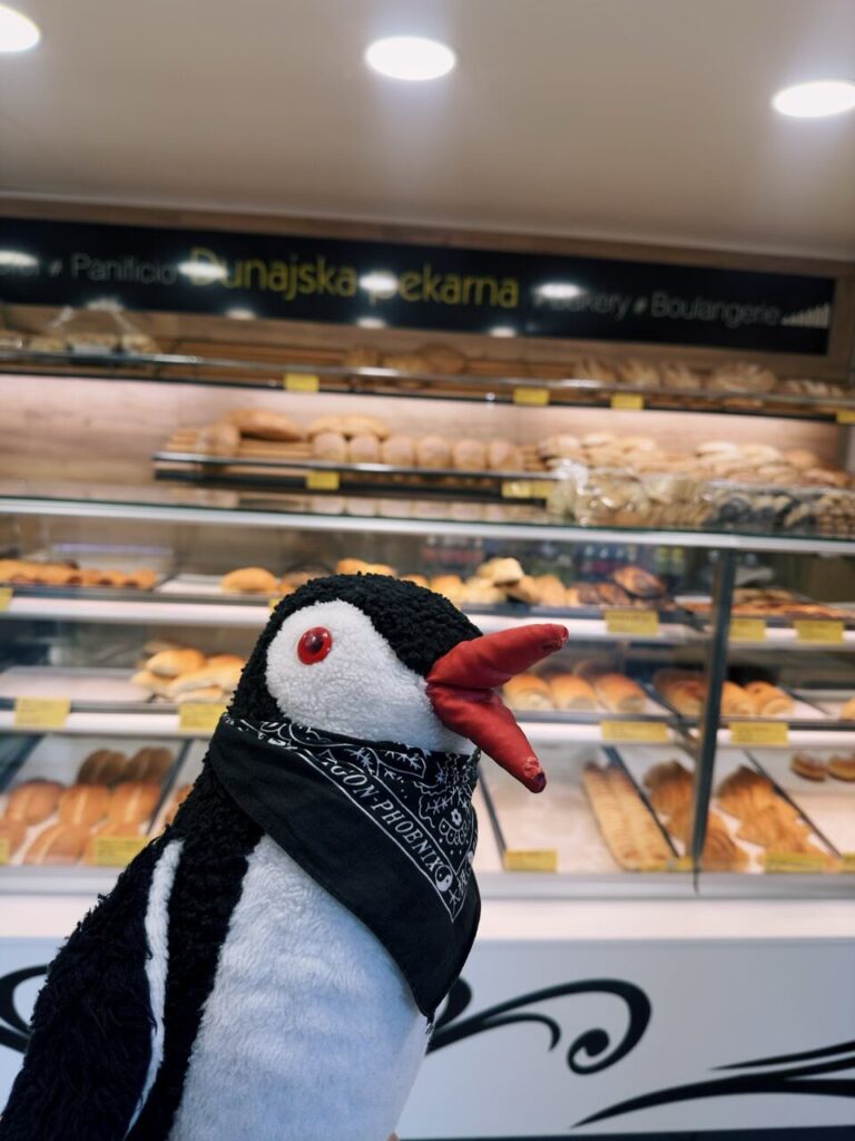 Plush penguin wearing a bandana stands before a display case of Slovenian bread