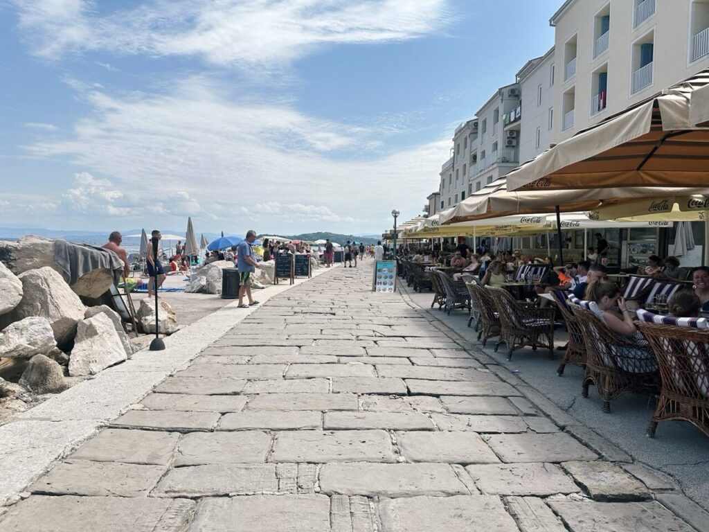 People sit at outdoor cafe tables along a stone promenade in Piran