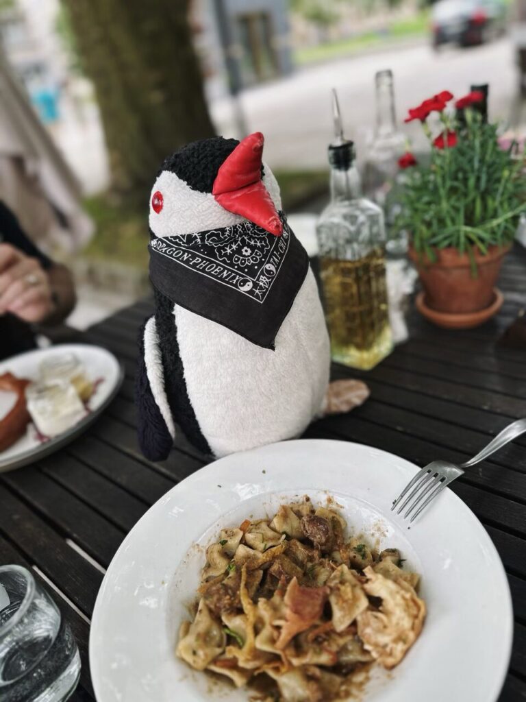 A plush penguin wearing a bandana sits at a table with pasta