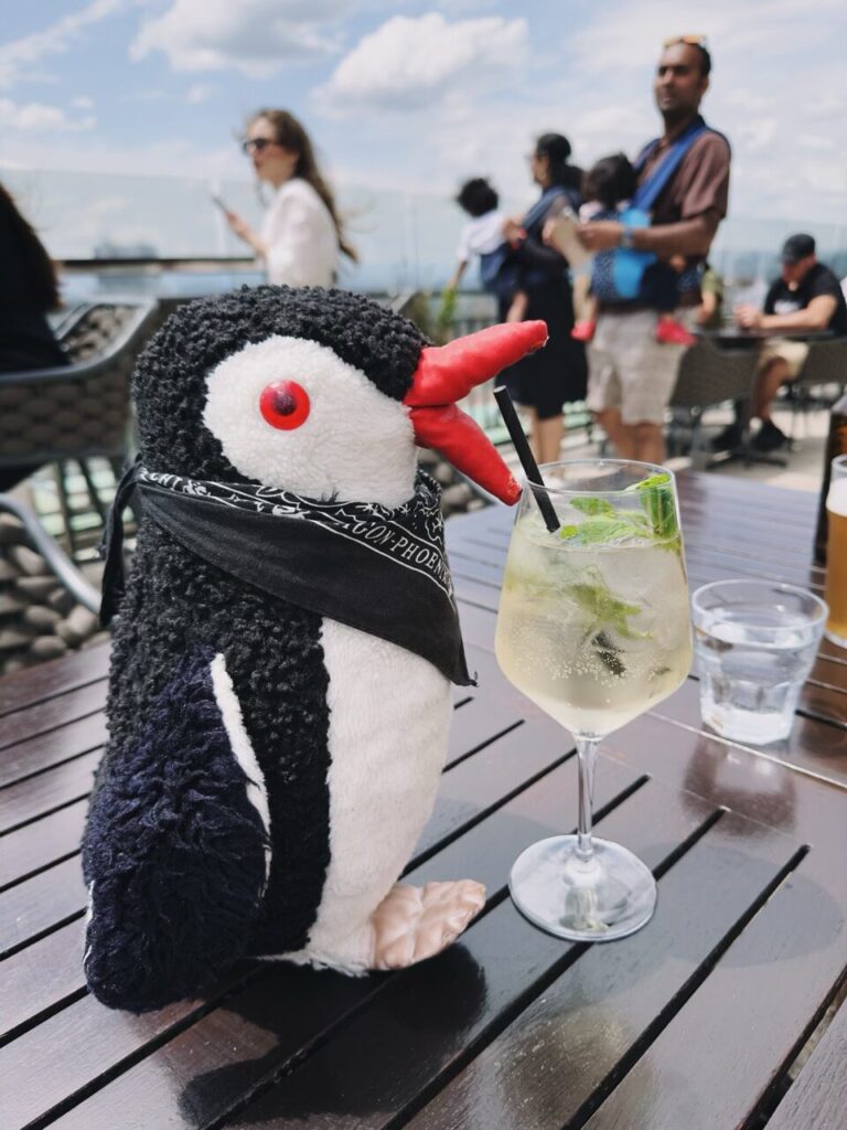 Plush penguin Miško wearing a bandana next to a cocktail at a restaurant in Slovenia