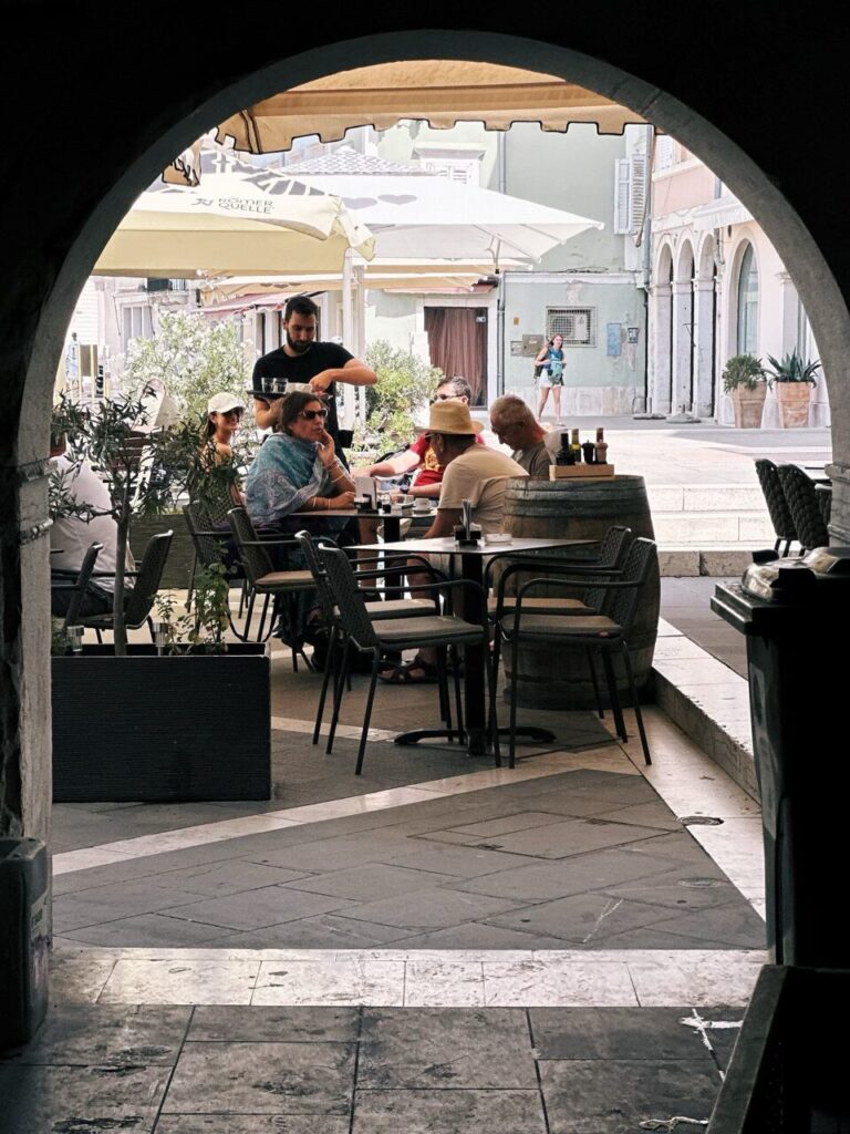 People dine at an outdoor cafe in Piran viewed through a stone archway