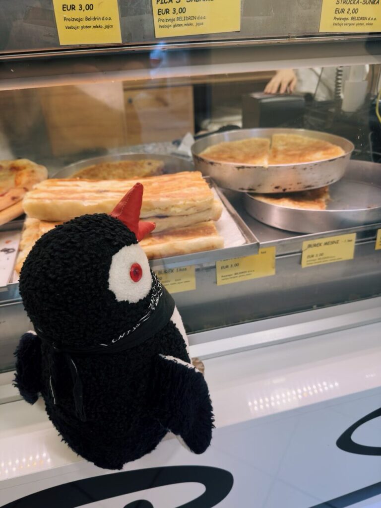 Plush penguin toy Miško looks at trays of burek in a Slovenia bakery display