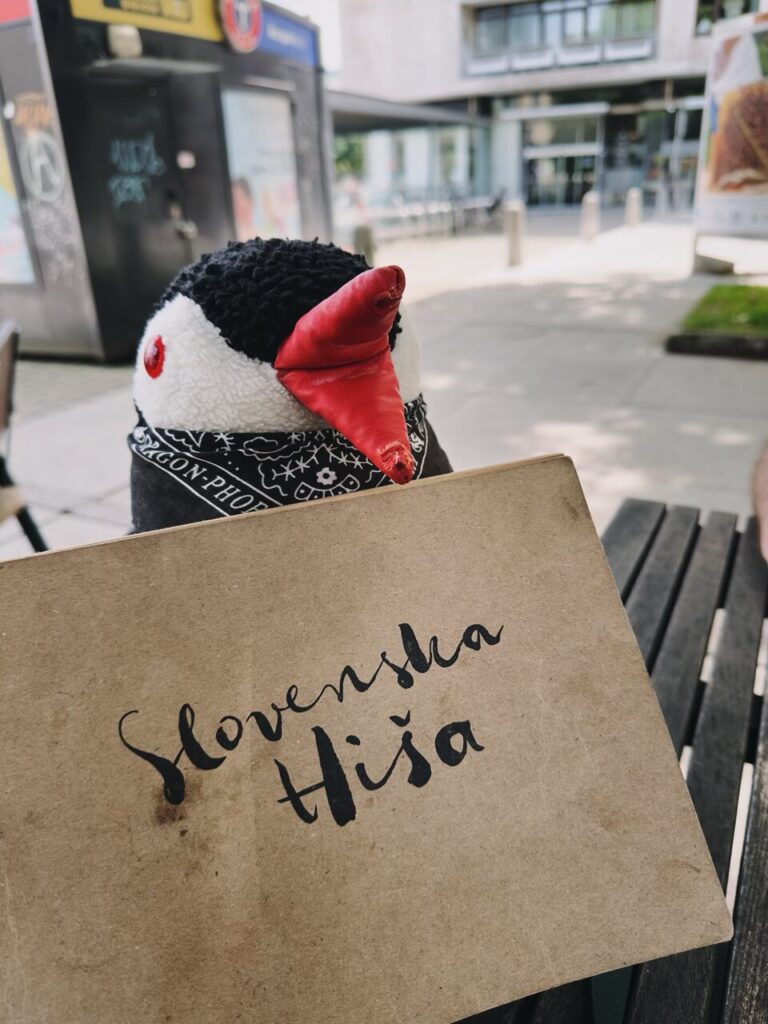 Plush penguin holds a menu labeled Slovenska Hiša at a restaurant in Slovenia