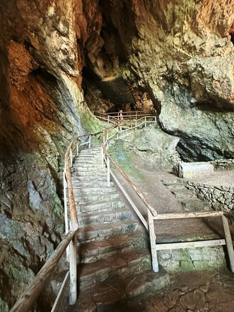 Stone stairs with wooden railings wind upward inside a large rocky cave