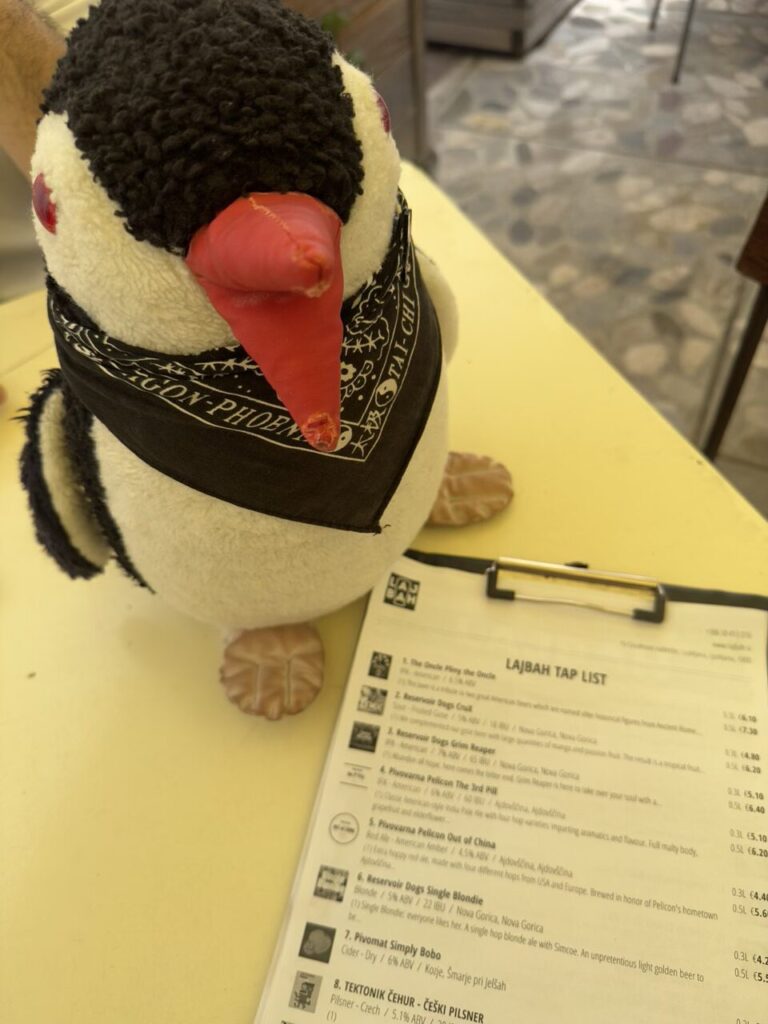 Plush penguin wearing a bandana sits next to a beer menu in Slovenia