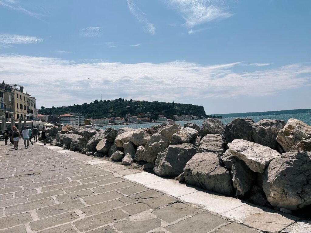 People walk along a stone promenade beside the sea in Piran, Slovenia