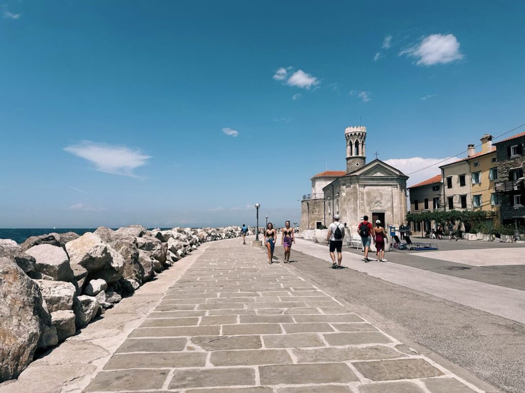 People walk along a stone promenade beside the sea in Piran