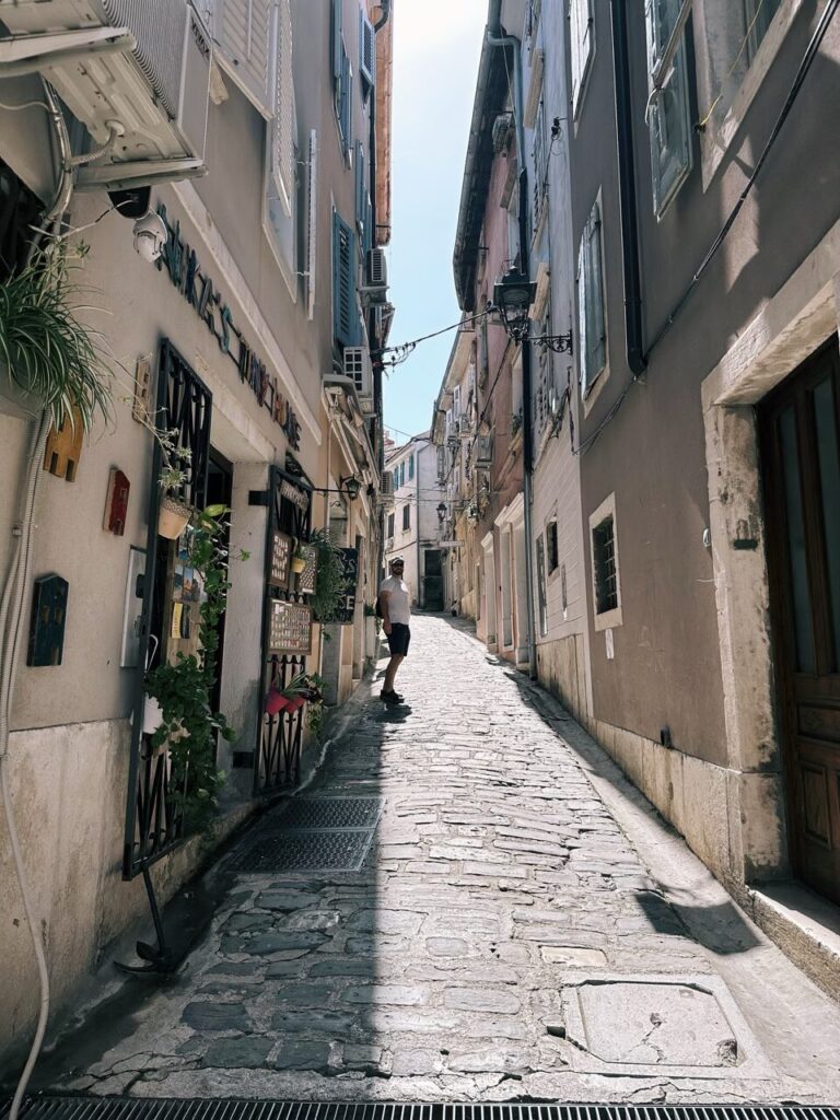 A man stands in a narrow, sunlit cobblestone alleyway in Piran