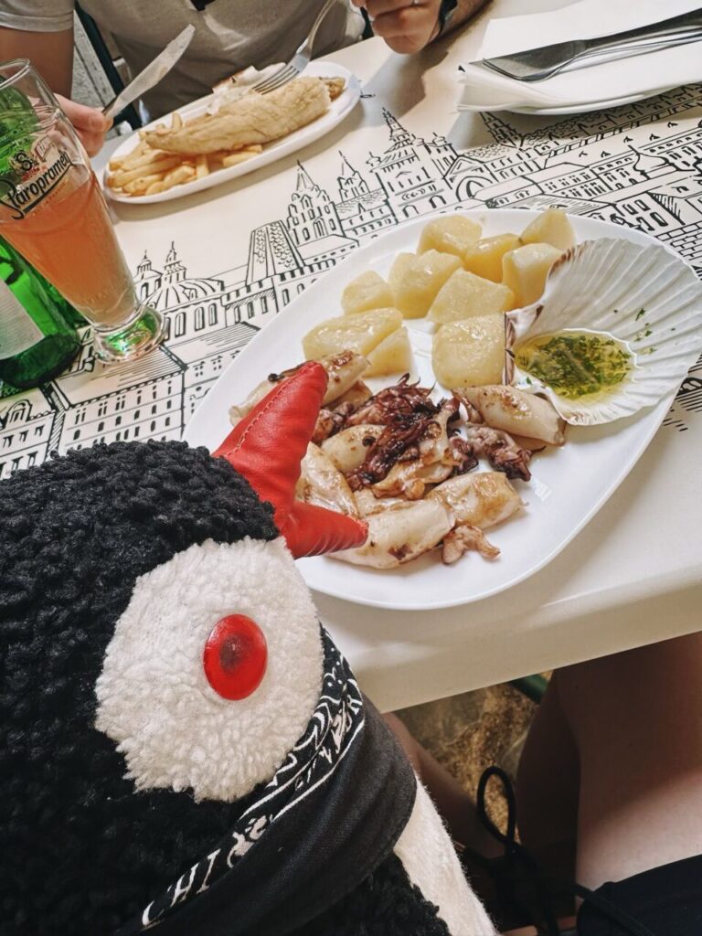 A plush toy sits at a table with grilled squid and potatoes