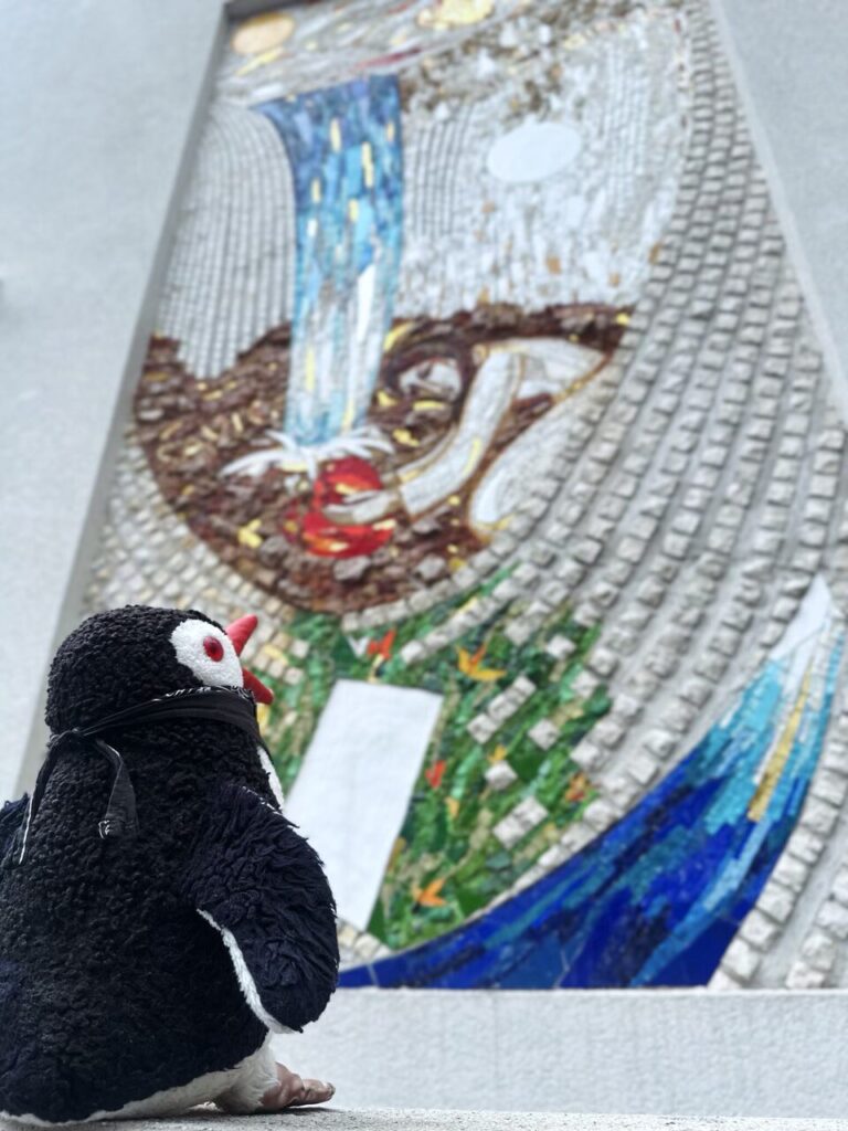 A plush penguin looks up at a colorful outdoor mosaic wall mural