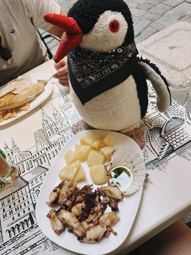 Plush penguin Miško sits at a Slovenia restaurant table with grilled squid and potatoes